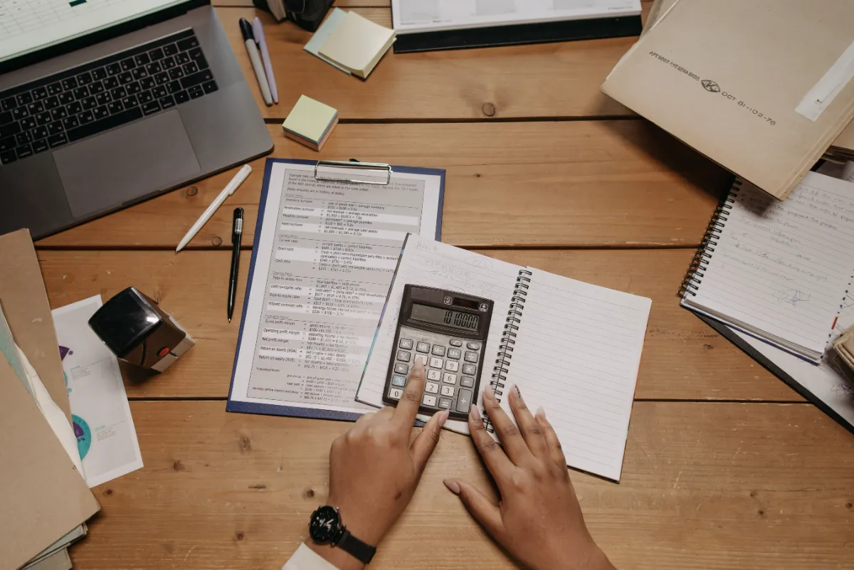 Vue de dessus d'un bureau en bois avec une calculatrice, des documents financiers et une personne en train de calculer